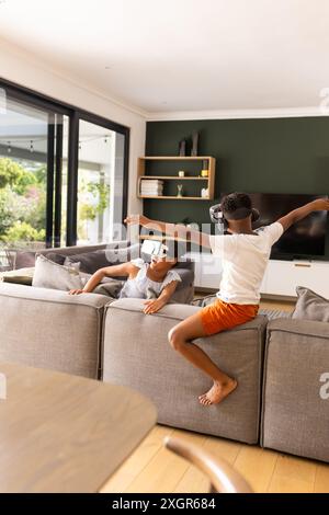 Fratello e sorella birazziali adolescenti apprezzano la realtà virtuale a casa. Sono immersi in un'esperienza di realtà virtuale in un accogliente salotto. Foto Stock