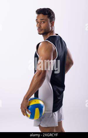 Il giovane uomo birazziale si pone con sicurezza nell'abbigliamento sportivo, tenendo una pallavolo su uno sfondo bianco. La sua posizione atletica suggerisce la preparazione per una competizione Foto Stock