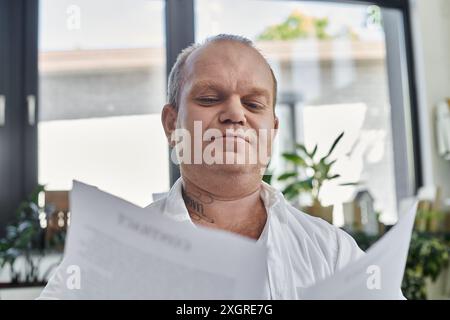 Un uomo con inclusività che indossa una camicia bianca siede in ufficio e legge i documenti. Foto Stock