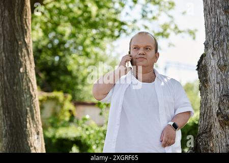 Un uomo con l'inclusività in una camicia bianca cammina in un parco, parlando al telefono. Foto Stock