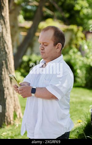 Un uomo con inclusività in una camicia bianca usa il telefono mentre cammina in un parco in una giornata di sole. Foto Stock