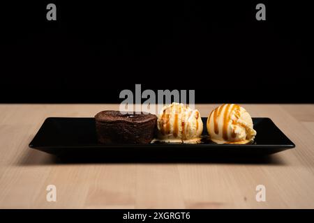 Un dessert di torta lavica al cioccolato e gelato alla vaniglia con gocciolamento di caramello su un piatto nero su un tavolo di legno su sfondo nero Foto Stock