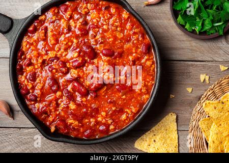 Chili con carne in vaschetta in ghisa con nachos su fondo in legno da vicino. Cucina messicana. Zuppa piccante con fagioli rossi, peperoni, pomodori e min Foto Stock