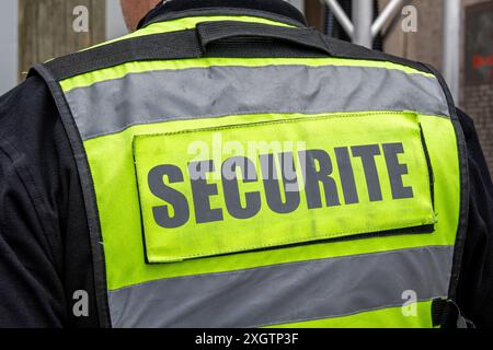 Gilet giallo fluorescente con la parola "sicurezza" scritta in francese sul retro e indossata da una guardia di sicurezza privata che guarda un sito sicuro Foto Stock