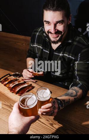 Un giovane allegro con i tatuaggi gode di un'esperienza culinaria informale in un ristorante barbecue, sorridendo mentre morde delle costolette succose e tiene un rinfresco Foto Stock