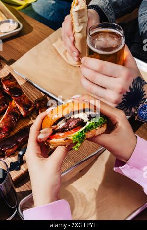 Una vivace scena di amici che condividono i pasti, tra cui hamburger, costolette e involtini, accompagnati da bevande fredde, a un tavolo in legno all'aperto durante un'informale ba Foto Stock