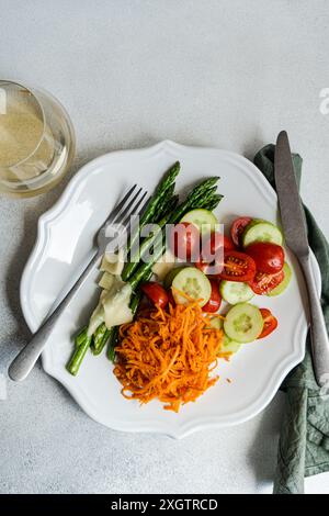 Dall'alto, un nutriente piatto di lance di asparagi, pomodori a fette, arrotondi di cetrioli e carote grattugiate con fette di formaggio. Un piatto bianco Foto Stock