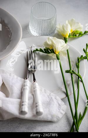 Un elegante tavolo con stoviglie in porcellana bianca, argenteria lucida avvolta in un tovagliolo di lino, un vetro testurizzato e delicati fiori freesia valorizzano Foto Stock