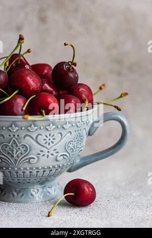 Ciliegie rosse appena lavate in una ciotola di ceramica blu ornata, con una ciliegia distesa all'esterno, posta su uno sfondo grigio testurizzato. Foto Stock