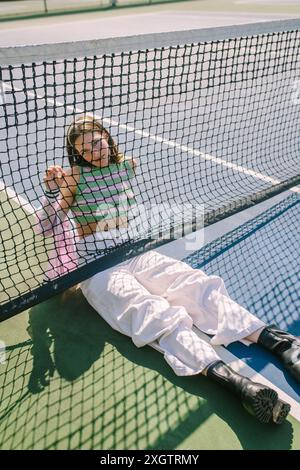 Una donna alla moda con un outfit alla moda, che include stivali neri e pantaloni bianchi, si rilassa contro una rete da tennis in una giornata di sole, l'immagine cattura una miscela Foto Stock