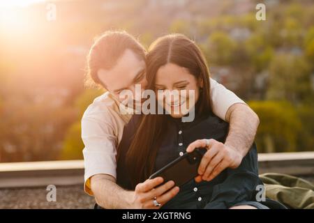 Una giovane coppia, un uomo e una donna, sono comodamente seduti insieme su un tetto durante un pomeriggio di sole. Entrambi sorridono mentre guardano un SM Foto Stock