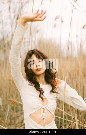 Una giovane donna con i capelli ricci si erge tra canne alte e asciutte, occhi chiusi e mani graziosamente sollevate, godendosi un momento di pace nella natura Foto Stock