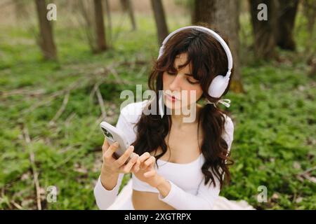 Una giovane donna con i capelli scuri ascolta la musica attraverso le cuffie bianche, sfogliando il suo smartphone in un ambiente lussureggiante e verdeggiante Foto Stock