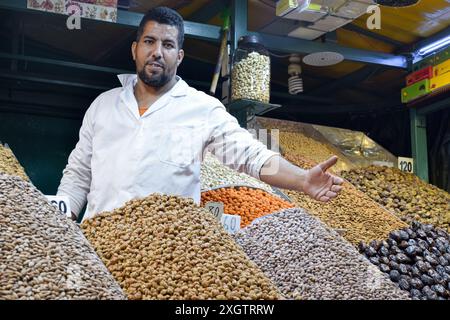 Un venditore maschile al mercato di Marrakech presenta un assortimento di frutta secca e noci. Sta guardando la telecamera e gestendo verso la merce con Foto Stock
