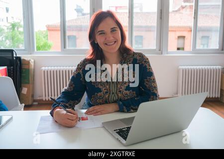 Una donna allegra e professionale si siede alla scrivania ben illuminata, sorridendo mentre lavora sul laptop e rivede i documenti Foto Stock