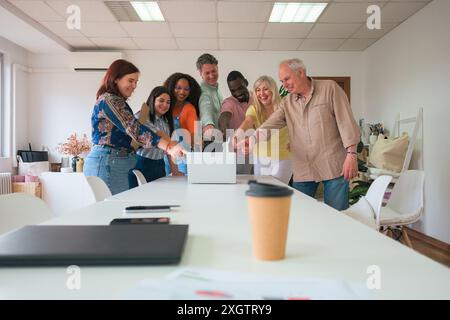 Un gruppo vivace di professionisti con background diversi si riunisce con entusiasmo intorno a un notebook in un ambiente luminoso per l'ufficio, puntando e condividendo i Foto Stock