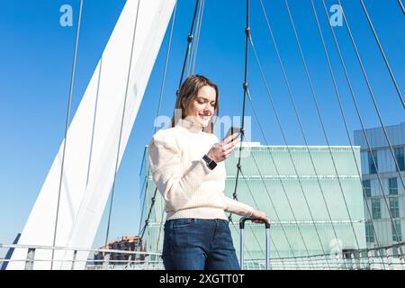 Una giovane donna che viaggia da sola sorride mentre usa il suo telefono cellulare su un ponte moderno con cieli limpidi Foto Stock