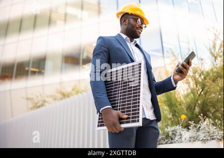 Dal basso uomo d'affari afroamericano con un vestito blu e un elmetto giallo, tenendo un pannello solare e uno smartphone all'aperto, guardando il suo telefono. Foto Stock