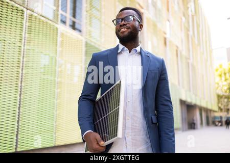 L'allegro uomo d'affari afroamericano, che guarda lontano dalla macchina fotografica, tiene un pannello solare mentre cammina lungo una strada soleggiata. Foto Stock