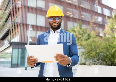 Un uomo d'affari afro-americano con tuta blu e elmetto giallo esamina i documenti mentre si trova fuori da un edificio urbano di uffici. Lui non guarda oltre fr Foto Stock