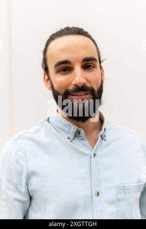 Un piacevole ritratto di un giovane uomo con barba folta e lunghi capelli legati alla schiena, vestito con una camicia in denim azzurro, sorridente dolcemente su una schiena semplice Foto Stock