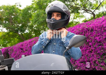 Un uomo con una giacca in denim che indossa un casco integrale nero con la sua moto, con vivaci fiori rosa sullo sfondo, che mostrano uno stile di vita di Foto Stock