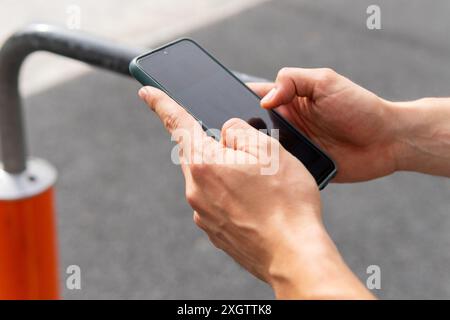 Vista ravvicinata anonima delle mani che gestiscono uno smartphone collegato a una stazione di allenamento in strada a Poblenou, evidenziando l'integrazione di technolo di oggi Foto Stock
