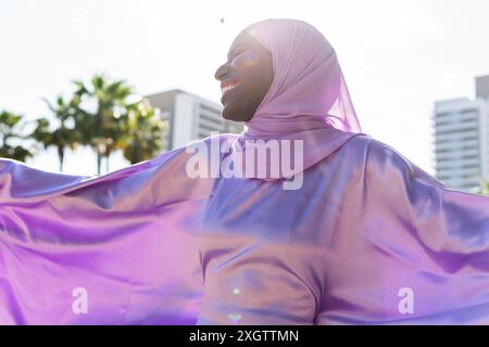 Una donna gioiosa che indossa un hijab di lavanda si staglia con le braccia allungate, crogiolandosi alla luce del sole, il suo sorriso irradia felicità, con uno sfondo urbano Foto Stock