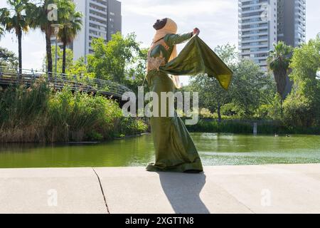 Una donna elegantemente vestita con un abito verde e un turbante si posa graziosamente in un tranquillo lago urbano circondato da lussureggiante vegetazione e moderni edifici Foto Stock