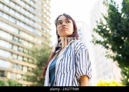 Una giovane donna con una camicia a righe e un paio di occhiali guarda in lontananza, catturata sullo sfondo di imponenti edifici urbani e vegetazione lussureggiante Foto Stock