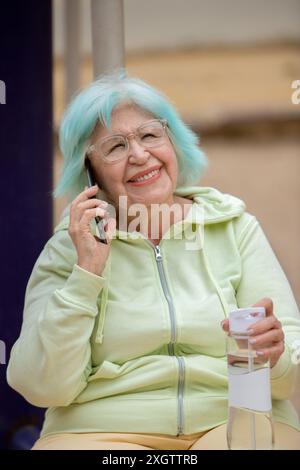 Donna anziana sorridente con capelli di tè unici utilizzando uno smartphone mentre è seduta nel parco. Ha in mano una bottiglia d'acqua e vestita con un abbigliamento sportivo, t Foto Stock