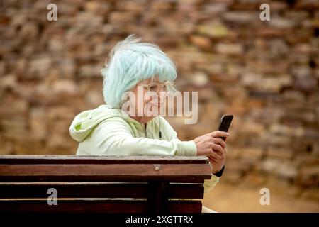 Una donna anziana con capelli blu vibranti sorride dolcemente mentre utilizza uno smartphone, seduta su una panchina del parco. È vestita casualmente con una felpa verde chiaro con cappuccio Foto Stock