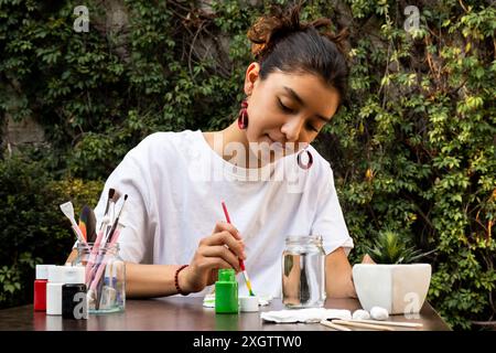 Una giovane donna concentrata ama dipingere oggetti di argilla all'aperto, circondati da una vegetazione lussureggiante. Dotata di vari colori di vernice e pennelli, realizza con intelligenza Foto Stock