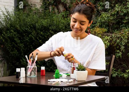 Giovane donna sorridente mentre dipinge decorazioni in argilla a un tavolo all'aperto, circondata da piante vibranti in un ambiente sereno giardino, mostrando la sua cr Foto Stock