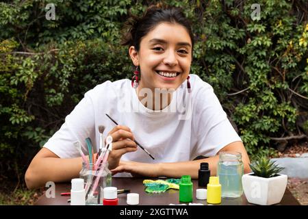 Una giovane e allegra donna si impegna con ansia a dipingere un colorato artigianato di argilla all'aperto, circondato da vasi di vernice brillante e natura Foto Stock