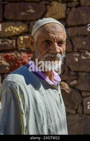 Un ritratto di un anziano marocchino che indossa abiti tradizionali e un berretto bianco, in piedi di fronte a un muro di pietra rustico in Nord Africa Foto Stock