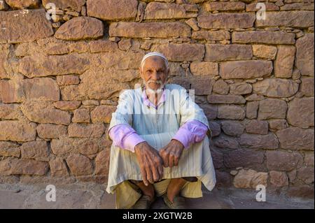 Un anziano marocchino siede contro un vecchio muro di pietra, vestito con abiti tradizionali marocchini, trasuda un senso di storia e patrimonio culturale Foto Stock