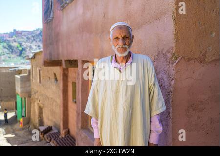 Un anziano uomo marocchino si erge con sicurezza davanti a una casa tradizionale del villaggio, indossando una classica djellaba bianca e una capsula, che incarna i ricchi c Foto Stock
