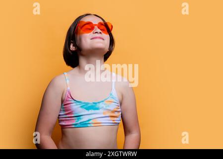 Una giovane ragazza che indossa occhiali da sole a forma di cuore e un costume da bagno, crogiolandosi alla luce del sole su uno sfondo arancione brillante Foto Stock