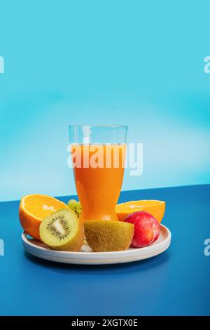 Una varietà di frutta fresca, tra cui arance, kiwi e un bicchiere di succo d'arancia disposti ad arte su un piatto, adagiato su un suggestivo sfondo blu. Foto Stock