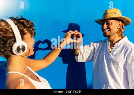 Un uomo con i dreadlock e un cappello e una donna con le cuffie formano una forma a cuore con le mani su uno sfondo blu che si guardano l'un l'altro Foto Stock