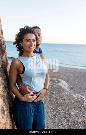 Una coppia caucasica vicino al mare. La donna, con una canotta bianca, guarda via con un sorriso brillante, mentre l'uomo con una camicia blu abbraccia dolcemente la h Foto Stock