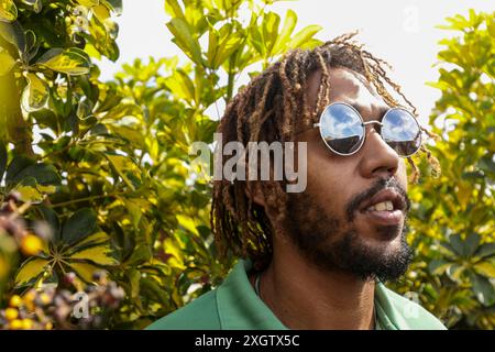 Un giovane afroamericano con una camicia verde, che indossa occhiali da sole riflettenti, guarda in lontananza tra un vivace fogliame verde. Foto Stock