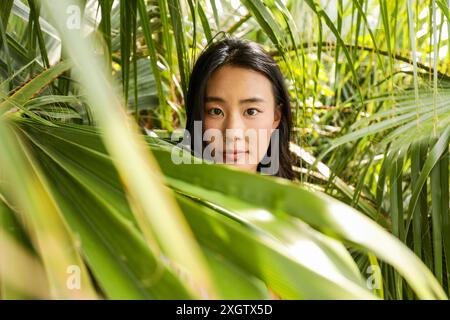 Una giovane donna asiatica guarda direttamente la macchina fotografica, il suo volto incorniciato da foglie tropicali verdi vibranti, trasmettendo un senso di curiosità e connessione con Foto Stock