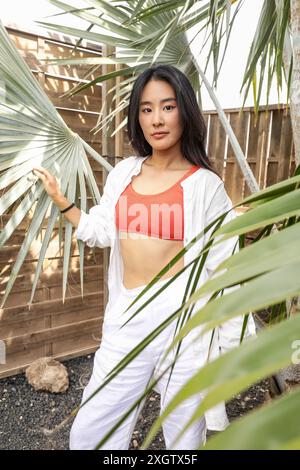 Una giovane donna asiatica che indossa una camicia bianca e una maglia rossa, guarda con sicurezza la fotocamera mentre si trova in piedi tra le lussureggianti fronde di palme verdi. Foto Stock