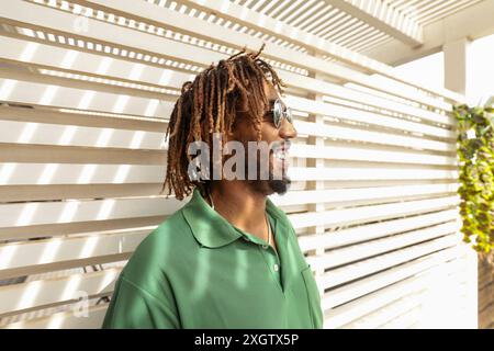 Un allegro afro-americano con paura e occhiali da sole e una polo verde ride di cuore, in piedi contro una schiena luminosa e a righe Foto Stock