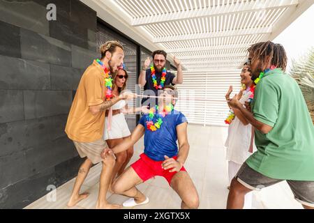 Gruppo multietnico di amici che giocano con entusiasmo a un gioco giocoso durante una riunione estiva, indossando colorate leis floreali ed esprimendo gioia. Foto Stock