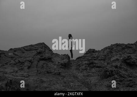 Una donna si staglia da sola sulle rocce aspre vicino al Faro di Favaritx a Minorca, catturata in un tono sereno e monocromatico Foto Stock