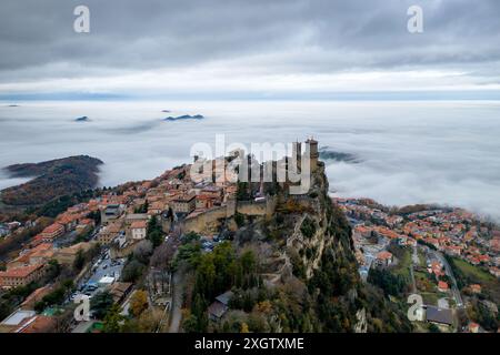 Una tranquilla vista aerea di San Marino mostra il paesaggio coperto di nuvole che circonda la prima Torre Guaita, un'iconica fortezza medievale, che sorge in primo piano Foto Stock