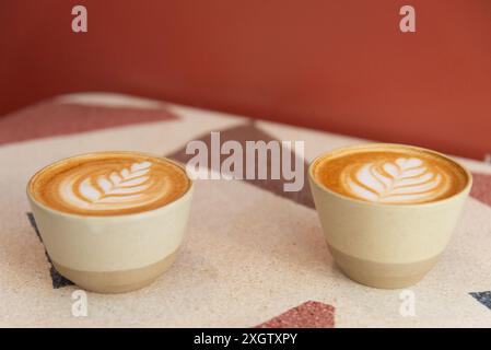 Questa immagine mostra due tazze di caffè con un'intricata latte art, posizionate fianco a fianco su un tavolo geometrico su uno sfondo rosso. Foto Stock